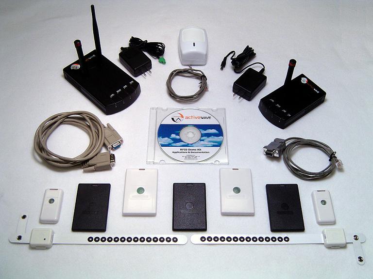 The Demo Kit laid out on a table: two black ActiveWave cases with antennas, a white motion detector, two power supplies, cables, a CD, and a row of tags, among them white and black card-shaped tags, two small white tags and two wristband tags.
